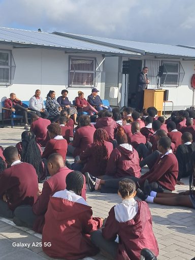 Students sitting on the ground listening to a speaker at a school event.