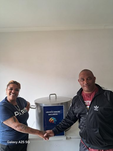 Two people shaking hands next to a large pot and a sign on the wall.