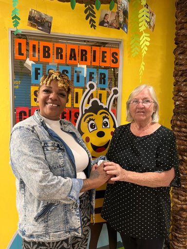 Two women smiling, standing beside a cheerful bee mascot in a vibrant library setting.