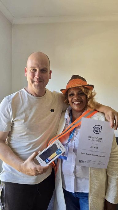 Two people smiling, one holding a certificate and a smartphone, wearing an orange hat.