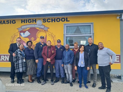 Group of people standing outside a school building with the name "Masig High School" visible.