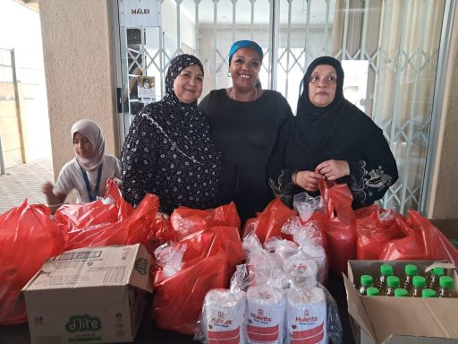Three women in headscarves stand beside bags of food donations, with a child in the background.